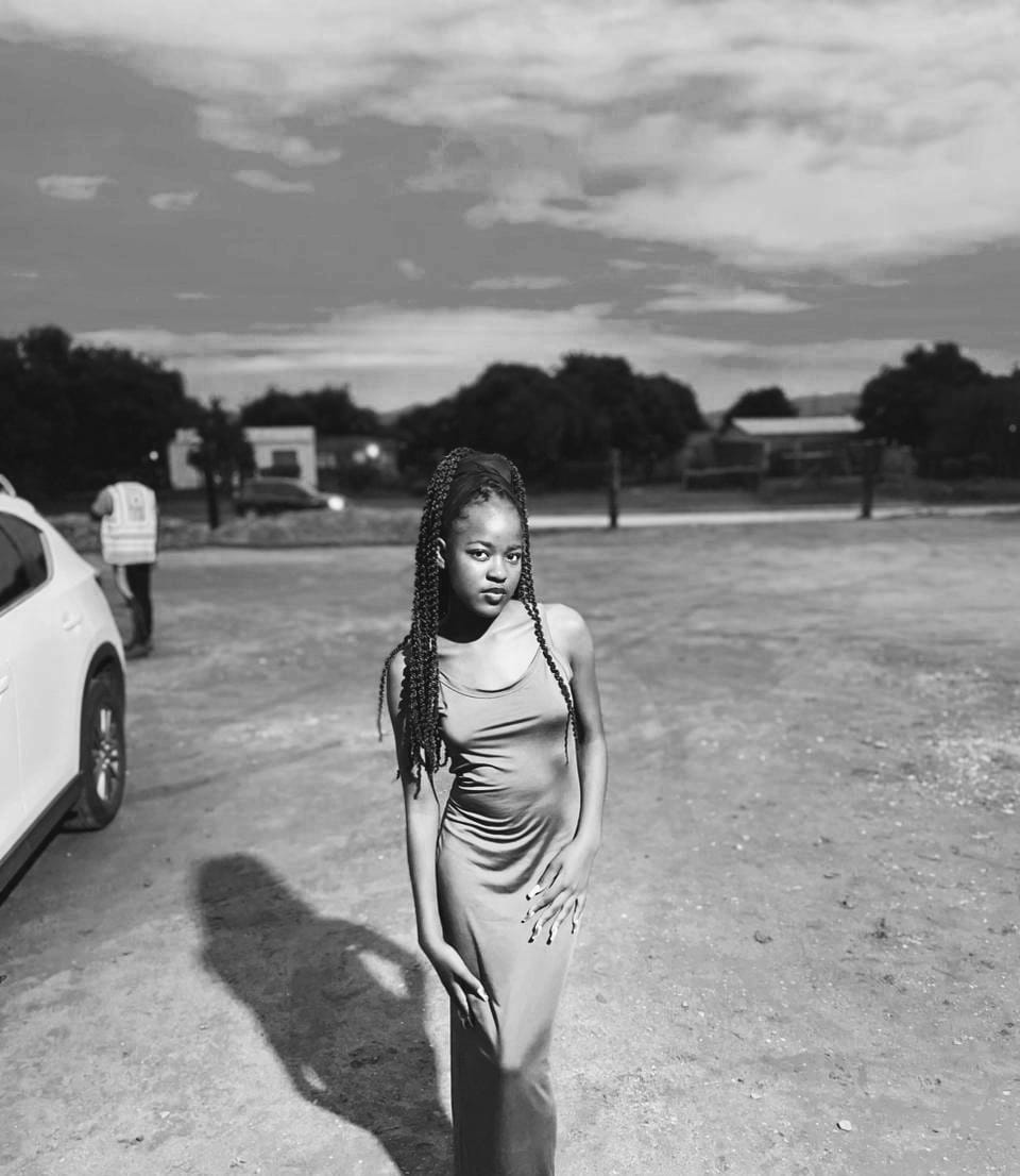 Black and white fashion portrait in a fitted dress, standing in an open dirt lot at dusk.