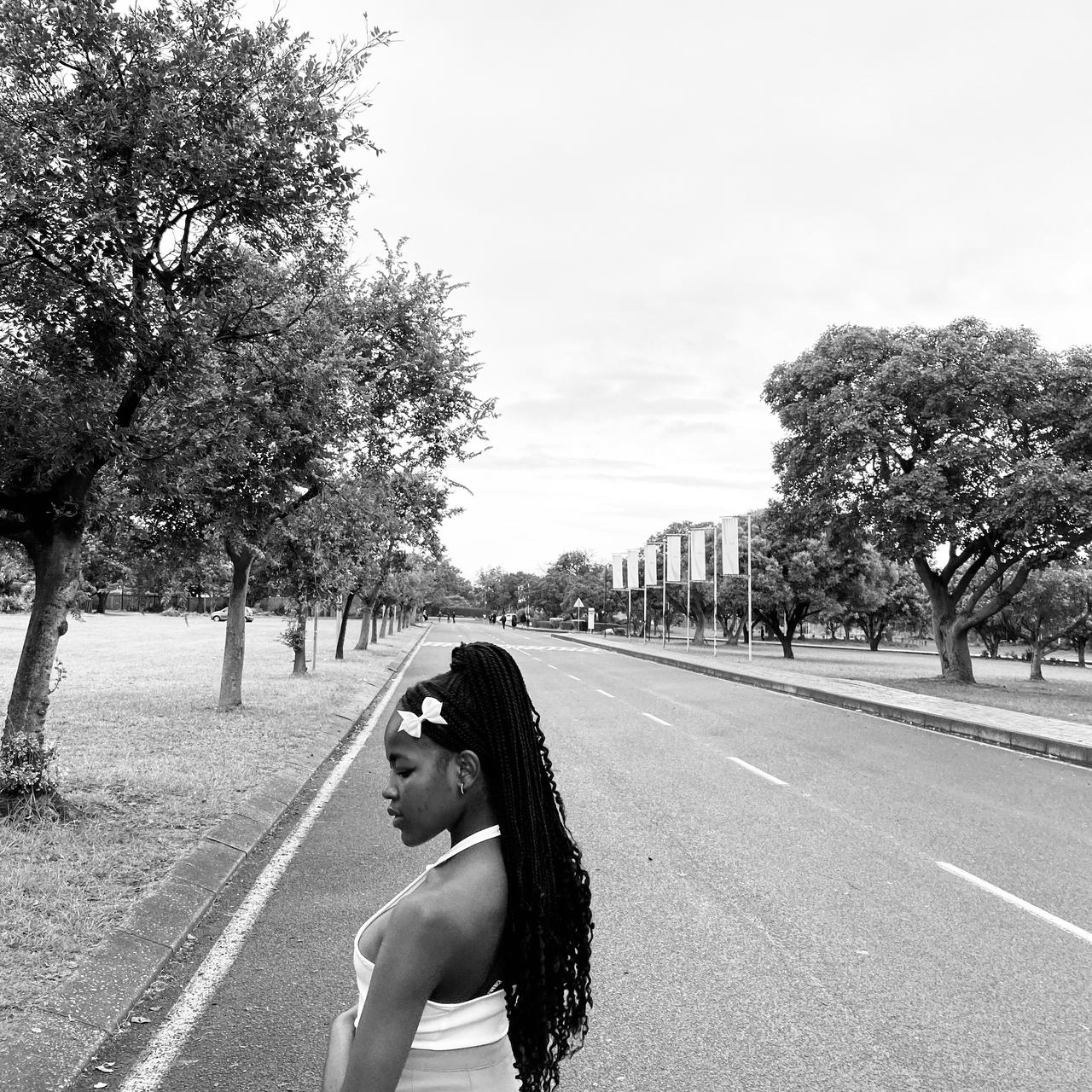 Black and white fashion portrait in profile on a tree-lined avenue, wearing a white halter top and braids.